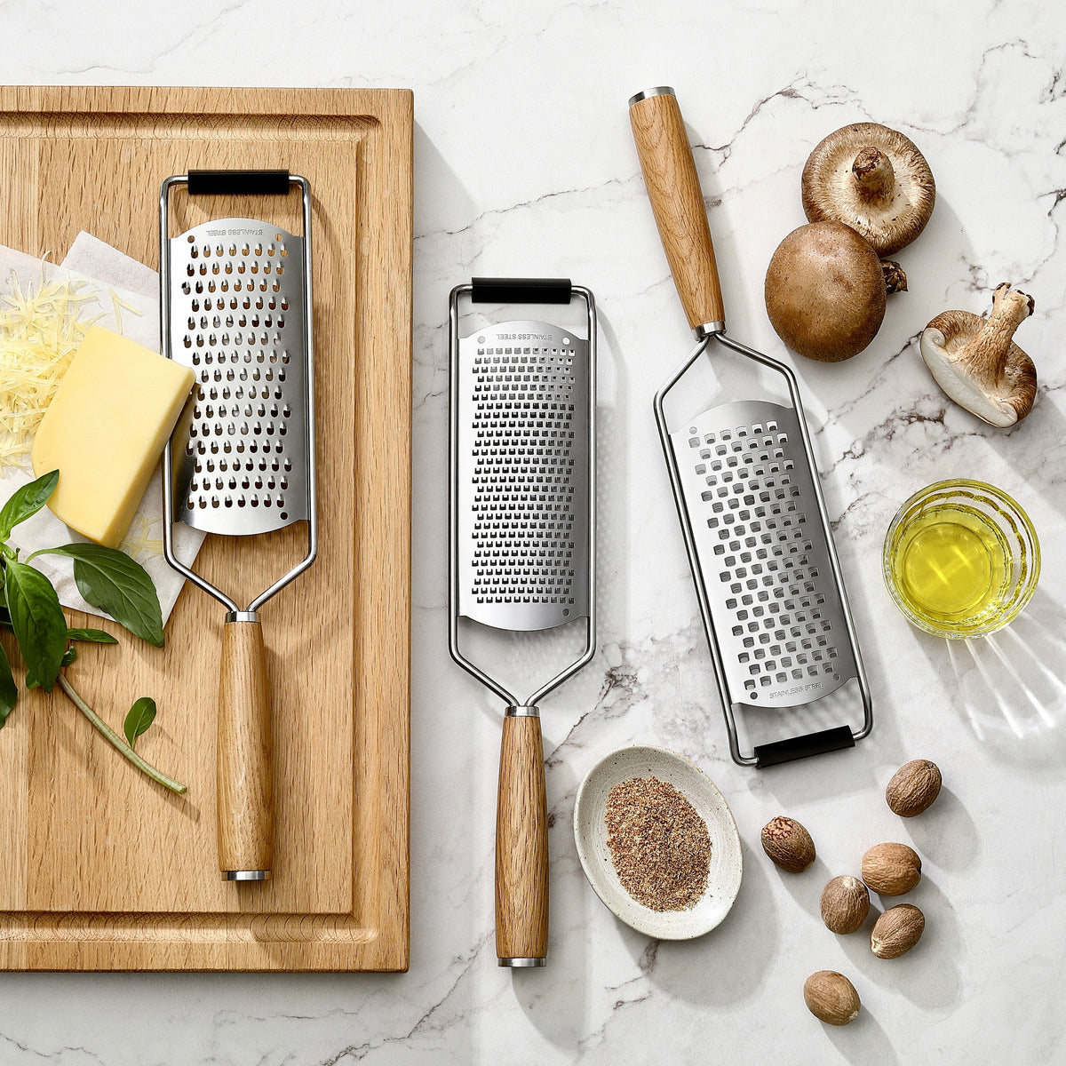 Feine, mittlere und grobe Reibe mit Griffen aus Eiche neben Käse, Muskatnuss und Pilzen