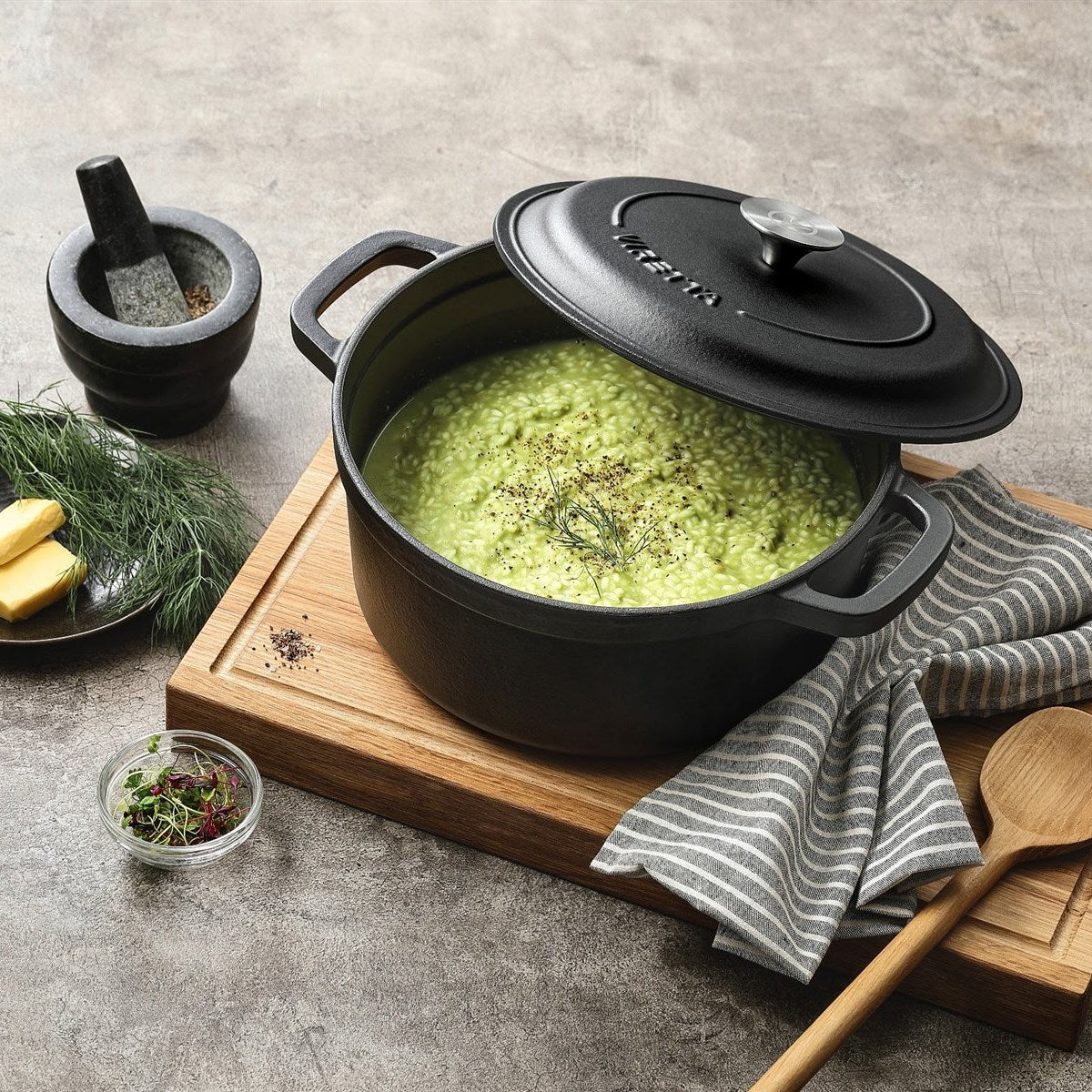 Schieferschwarzer Gusseisentopf mit grünem Risotto und schräg aufgelegtem Deckel, auf einem Schneidebrett #Farbe_Schieferschwarz