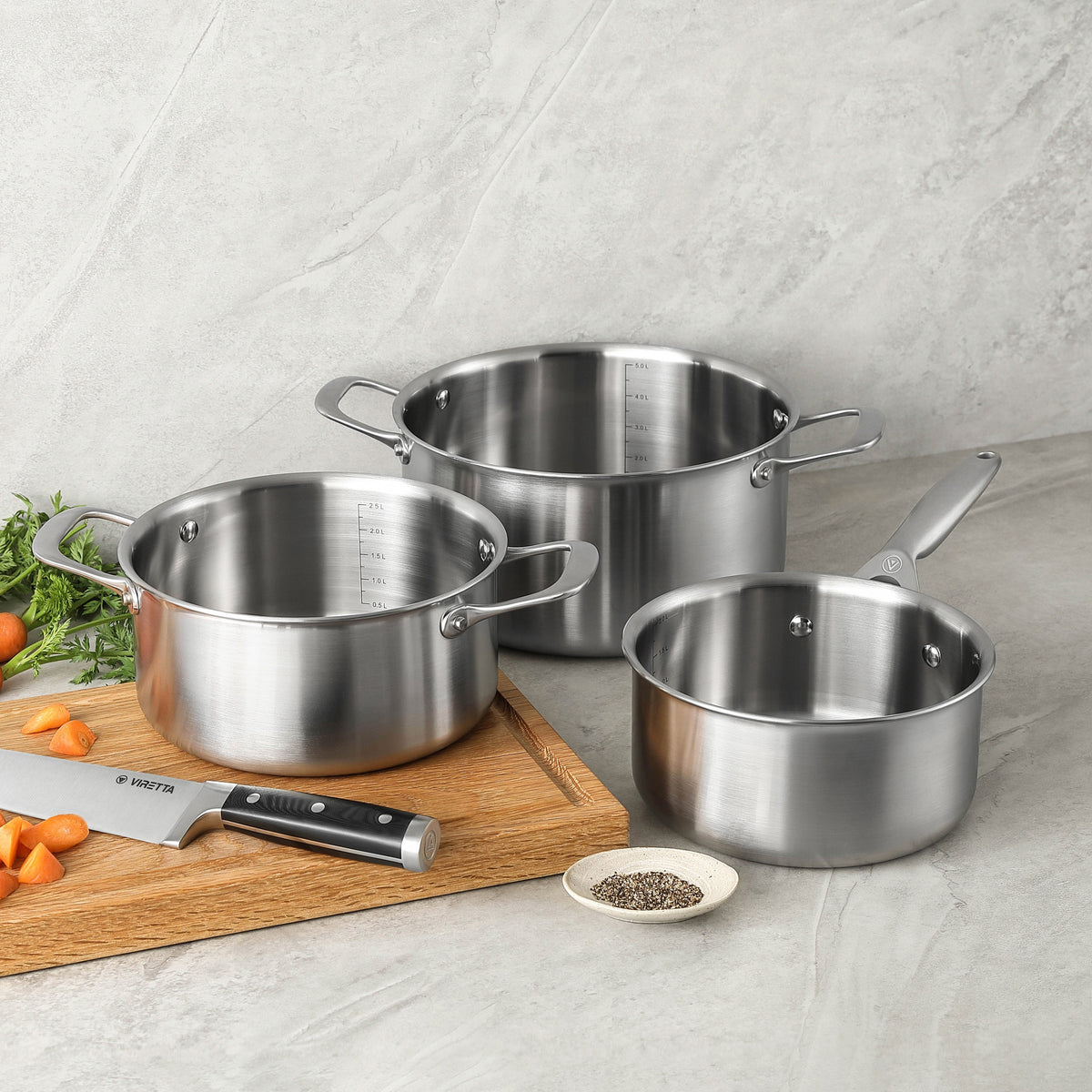 Kasserolle und Kochtöpfe aus 3-Ply Edelstahl nebeneinander auf grauem Stein und einem Schneidebrett