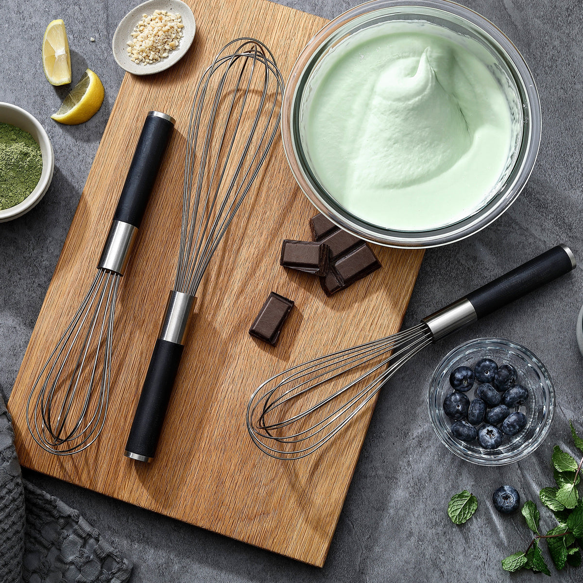 Drei Schneebesen mit Griffen aus G-10 neben einer Schüssel mit grüner Mousse und Schokolade