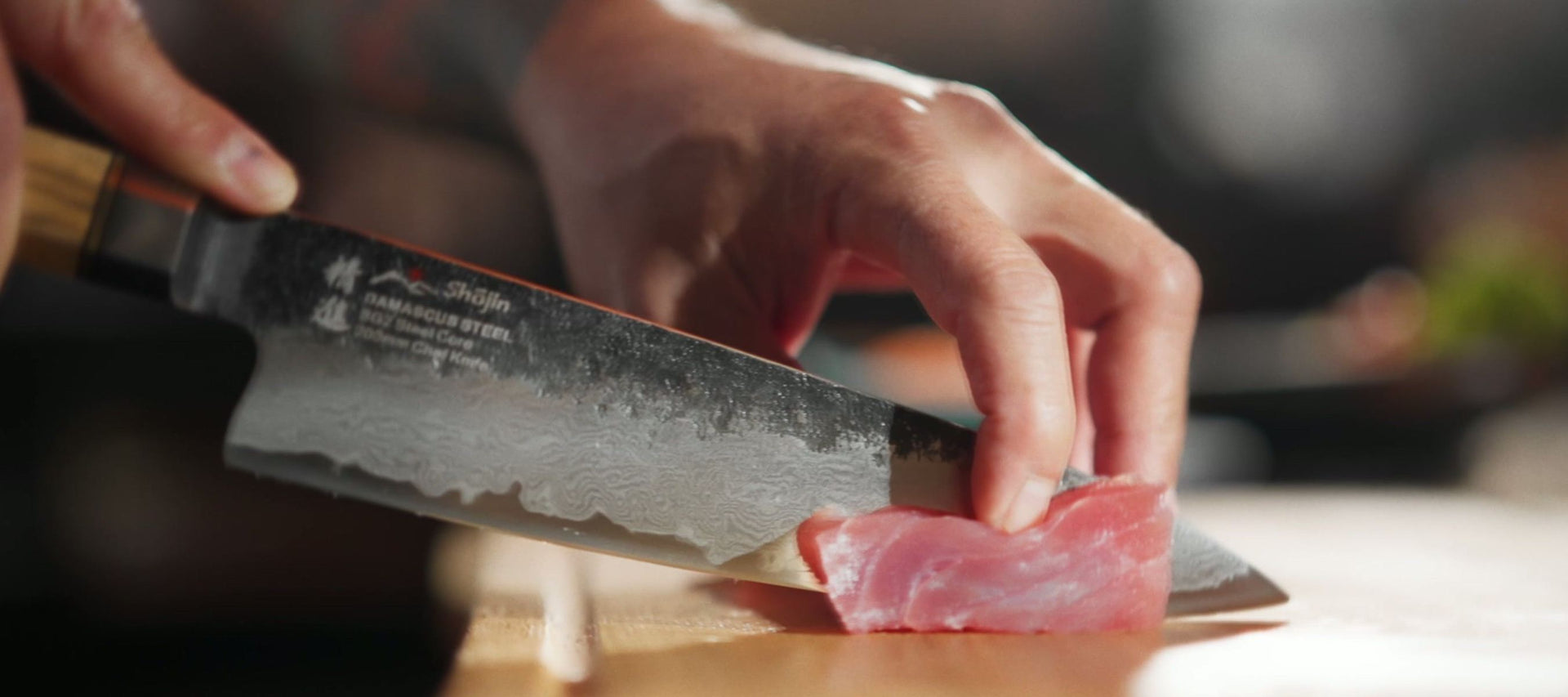 Eine Hand schneidet ein Stück Thunfisch mit einem Shōjin-Messer mit Damastmuster auf einem Schneidebrett aus Holz