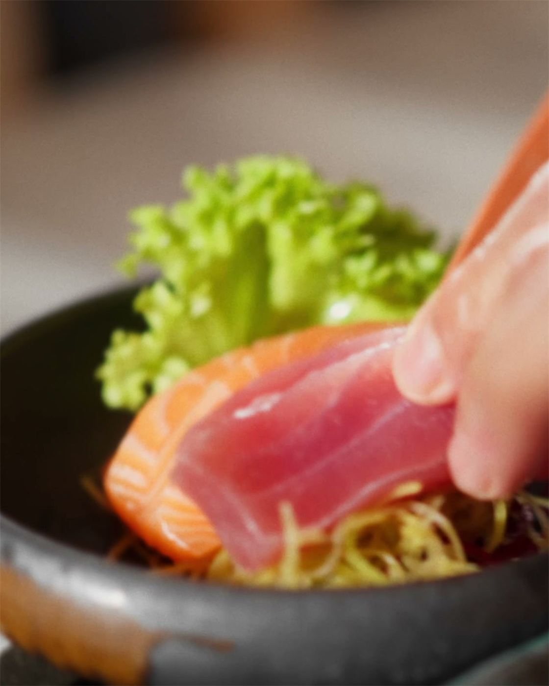 Eine Hand legt ein Stück Thunfisch auf einen dunklen Teller mit Lachs und Salat