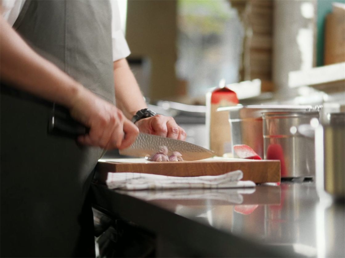 Ein Koch mit grauer Schürze hackt Schalotten mit einem Messer mit gehämmerter Klinge auf einem Schneidebrett in einer Restaurantküche