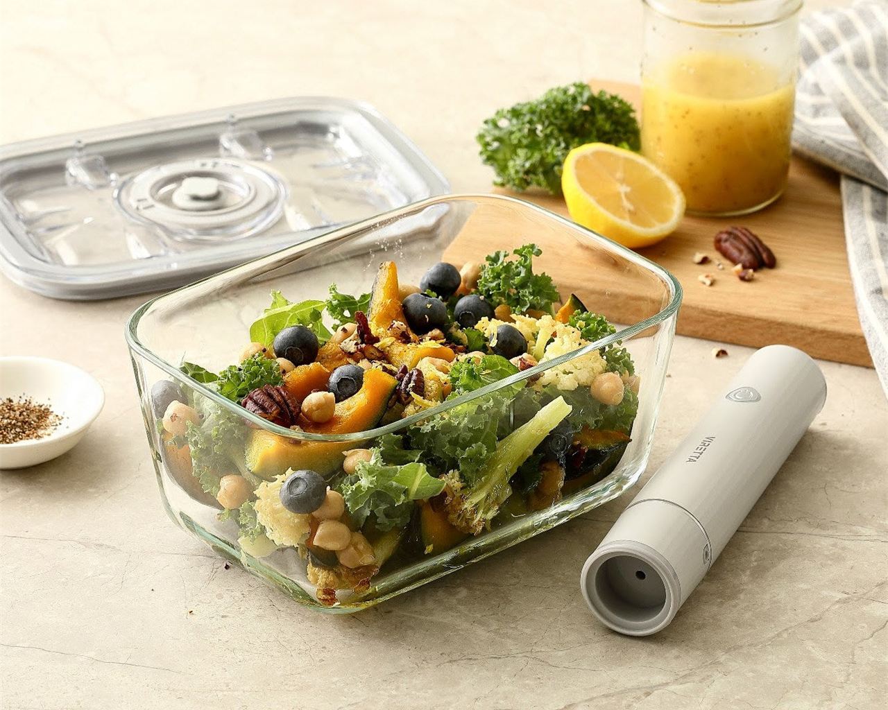 Ein Vakuumbehälter aus Glas mit Salat; der Deckel und die elektrische Vakuumpumpe liegen daneben