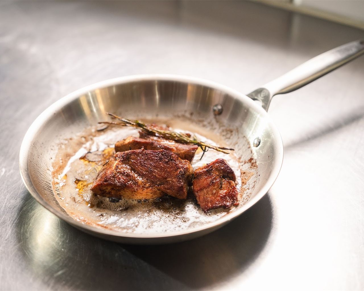 Eine Bratpfanne aus Edelstahl mit gebratenem Fleisch und einem Kräuterzweig auf einer Arbeitsfläche aus Stahl
